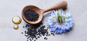 Black nigella seeds in a wooden spoon next to a blue nigella flower on a textured gray background.