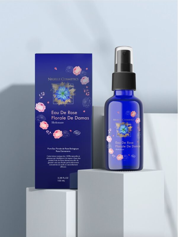 Flacon spray d'eau de rose bio avec des motifs floraux posé à côté de sa boîte sur un support géométrique et un fond bleu clair.