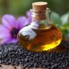 Flacon d'huile de nigelle entouré de graines noires et de fleurs de Nigella sativa